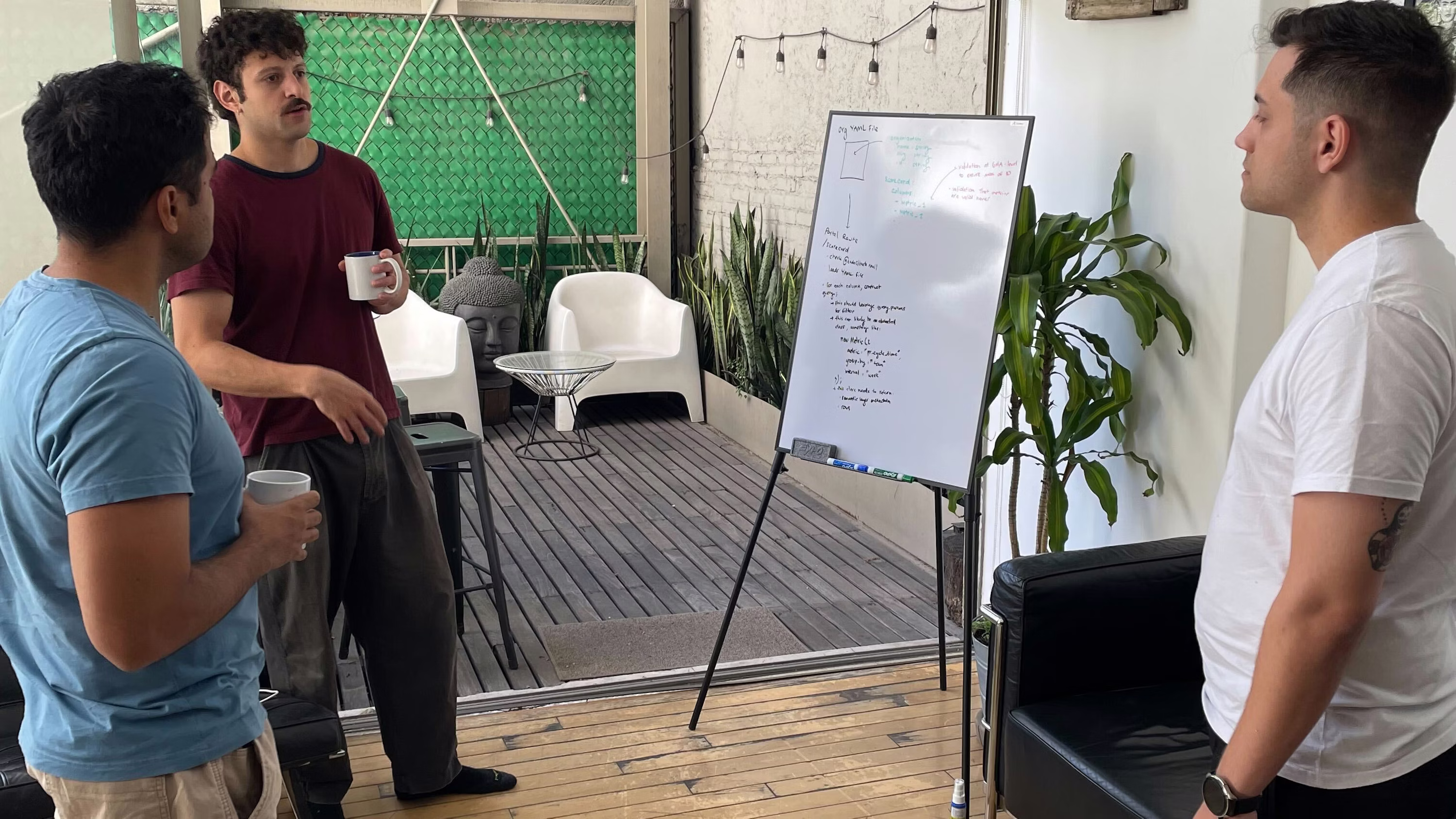 Three men having a discussion while holding coffee mugs around a whiteboard with notes in a modern indoor-outdoor space.