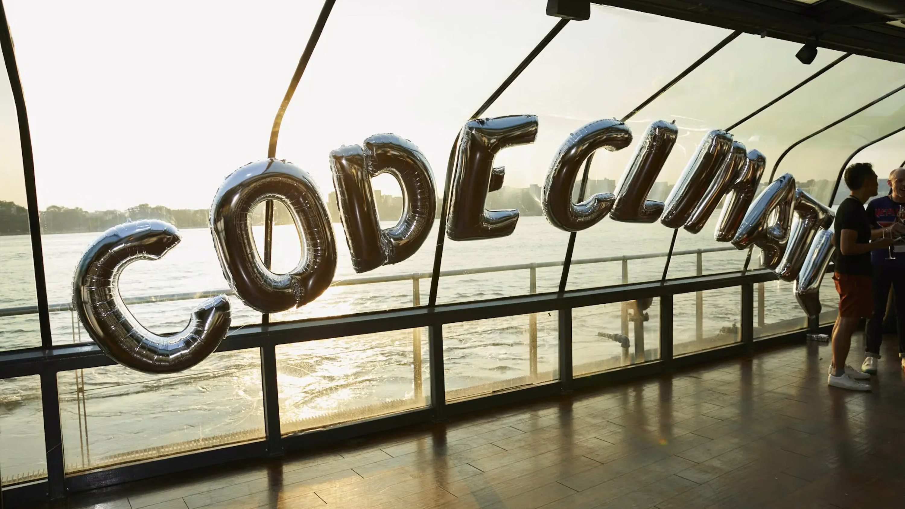 Silver foil balloons spelling 'CODE CLIMATE' hanging in front of a large glass window overlooking water with people talking nearby.