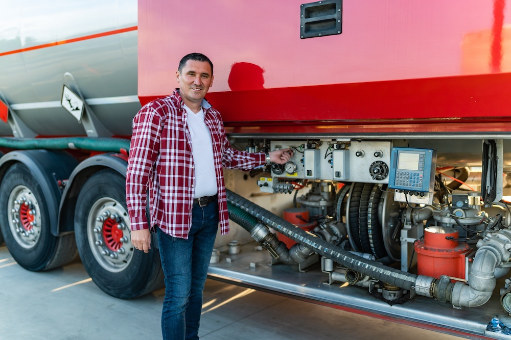 Man in plaid shirt inspecting fuel and mechanical controls on a red tanker truck, ensuring vehicle safety and maintenance compliance