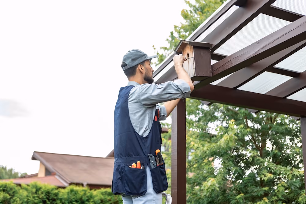 Handyman installing a wooden birdhouse on backyard pergola with tool belt and cap on