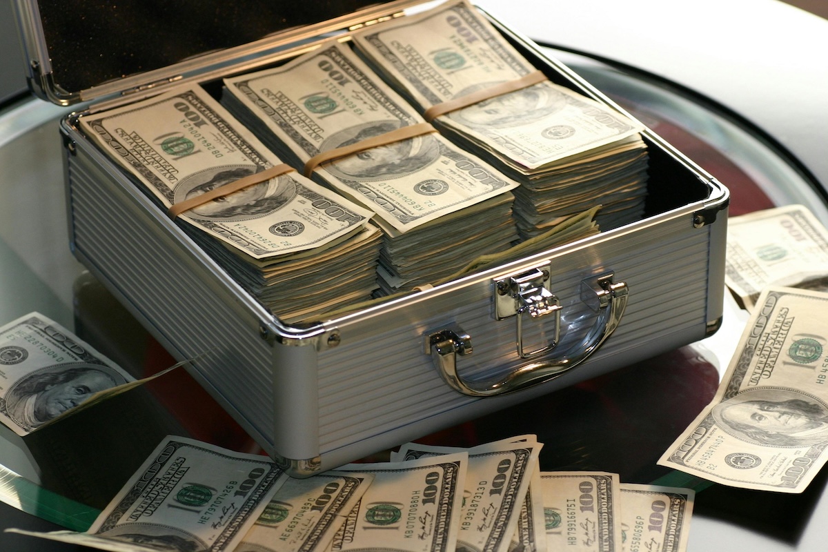 A small meta hand case full of perfectly stacked cash sits open on a table by loose stacks of cash in front of it.