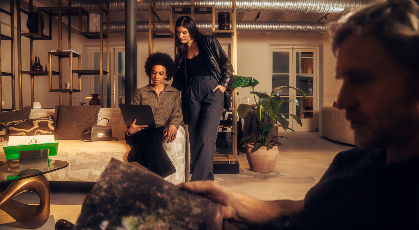 Two women in a modern living room, one sitting on a couch using a laptop and the other standing next to her, while a man is in the foreground reading a magazine.