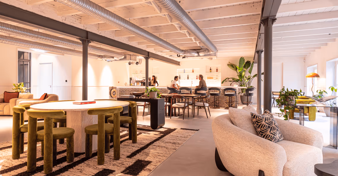 Spacious modern living and dining area with round dining table, beige sofas, bar stools, and indoor plants under exposed ceiling beams.