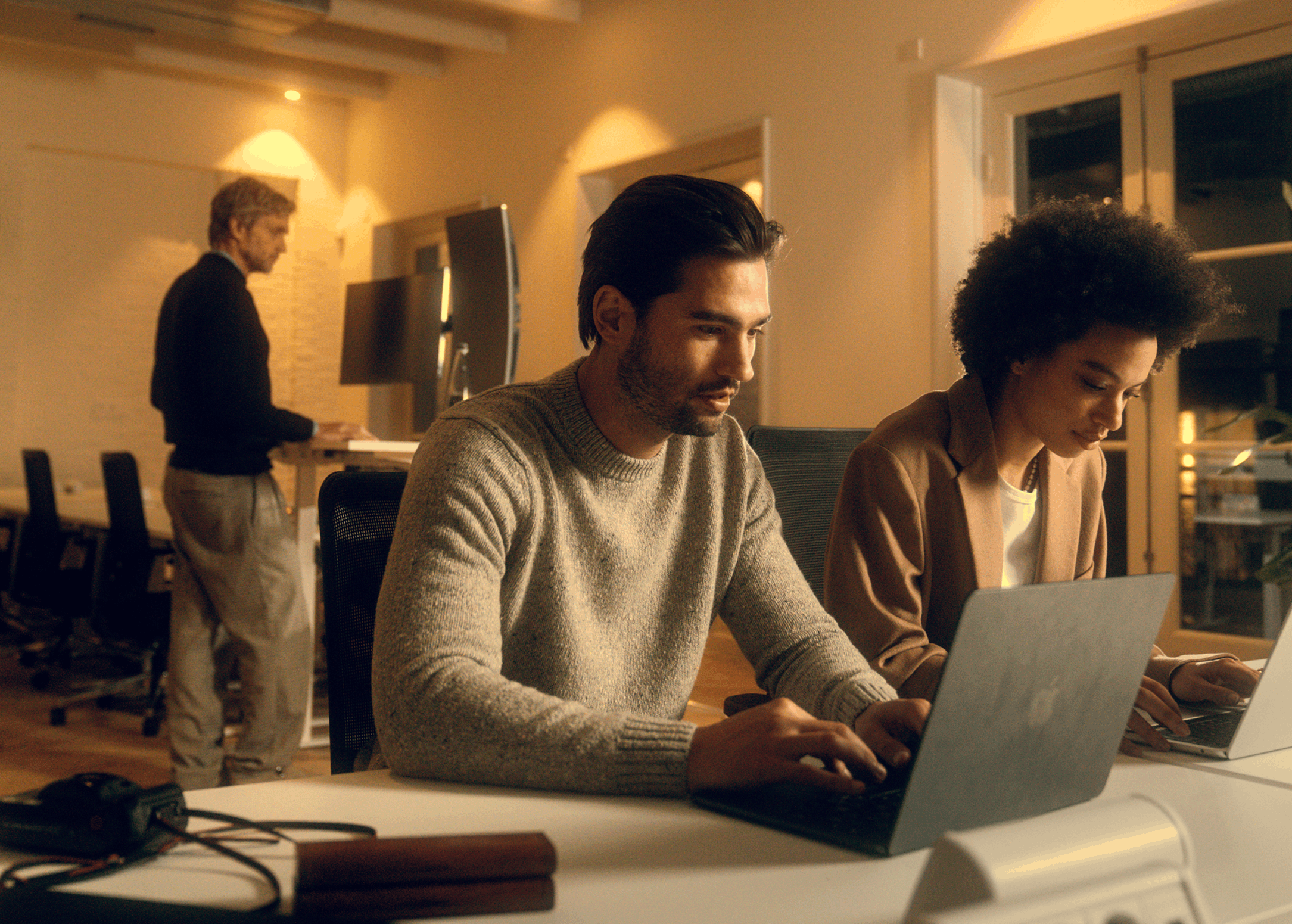 Two young professionals working on laptops at a desk in a warmly lit office, while a third person stands working in the background.