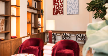 Modern cozy seating area with two red velvet chairs, a white side table, decorative shelves, and abstract wall art.
