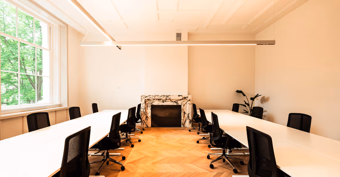 Bright modern conference room with two long white tables, black swivel chairs, a marble fireplace, wooden floor, and a large window showing green trees.