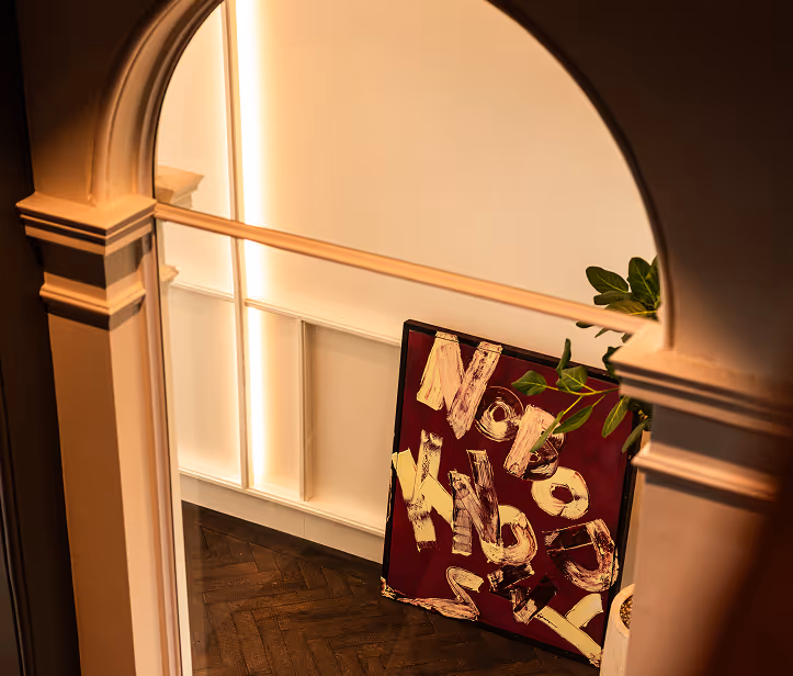 Framed abstract painting with bold letters leaning against a wall seen through an arched doorway.