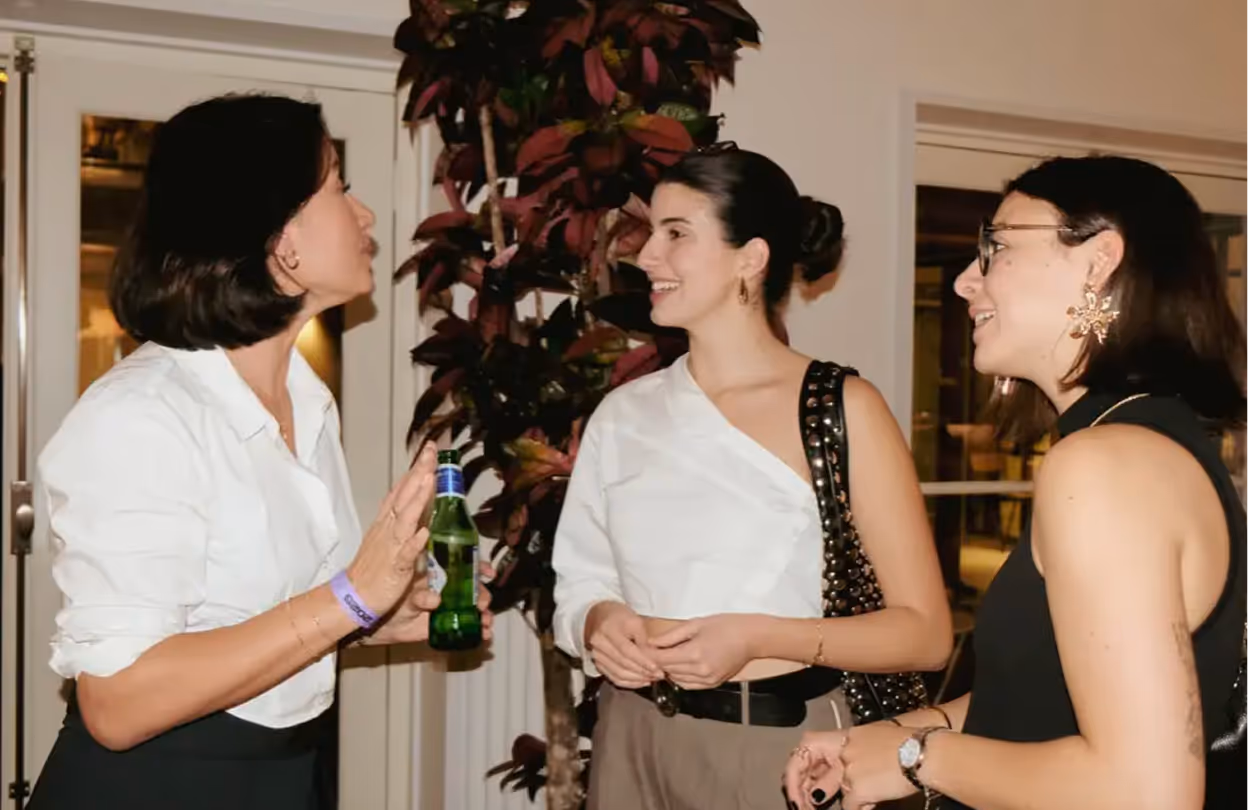 Two young men smiling and chatting at a bar or social event with blurred people in the background.