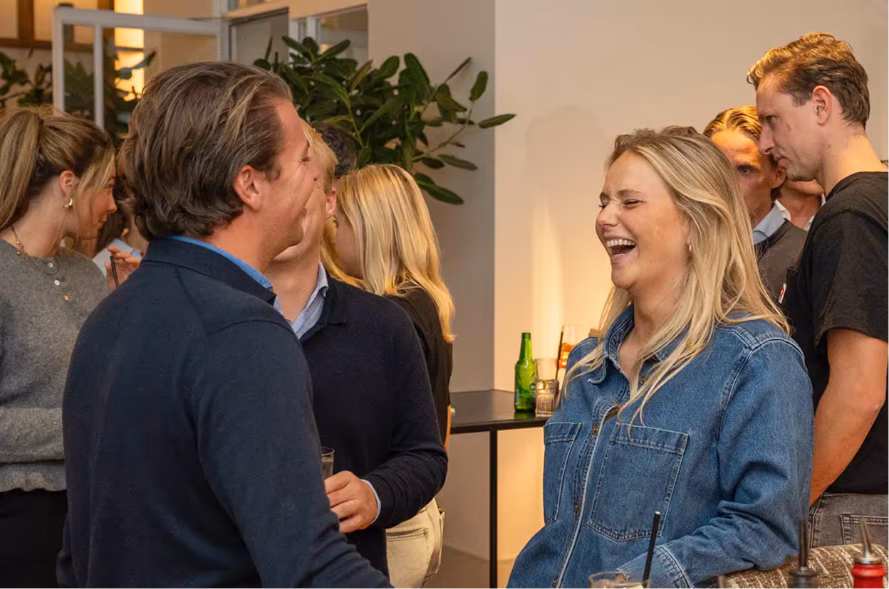 Group of people socializing indoors, a woman in a denim jacket laughing while talking to a man in a dark sweater.