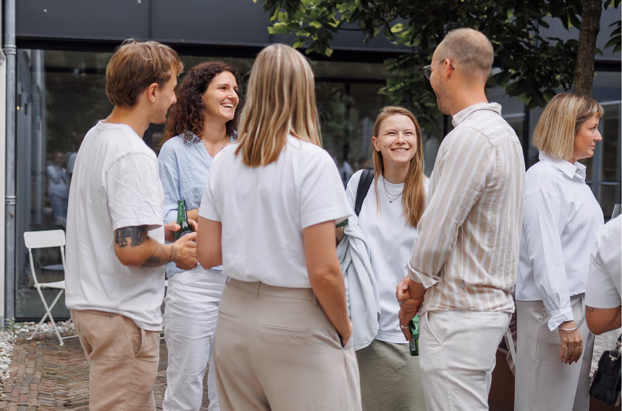 Group of six people standing outdoors, smiling and conversing, some holding drinks.