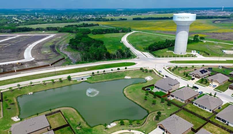 Aerial view of a residential neighborhood and water tower in Princeton, Texas, featuring new housing developments, green fields, and a small pond with a fountain.