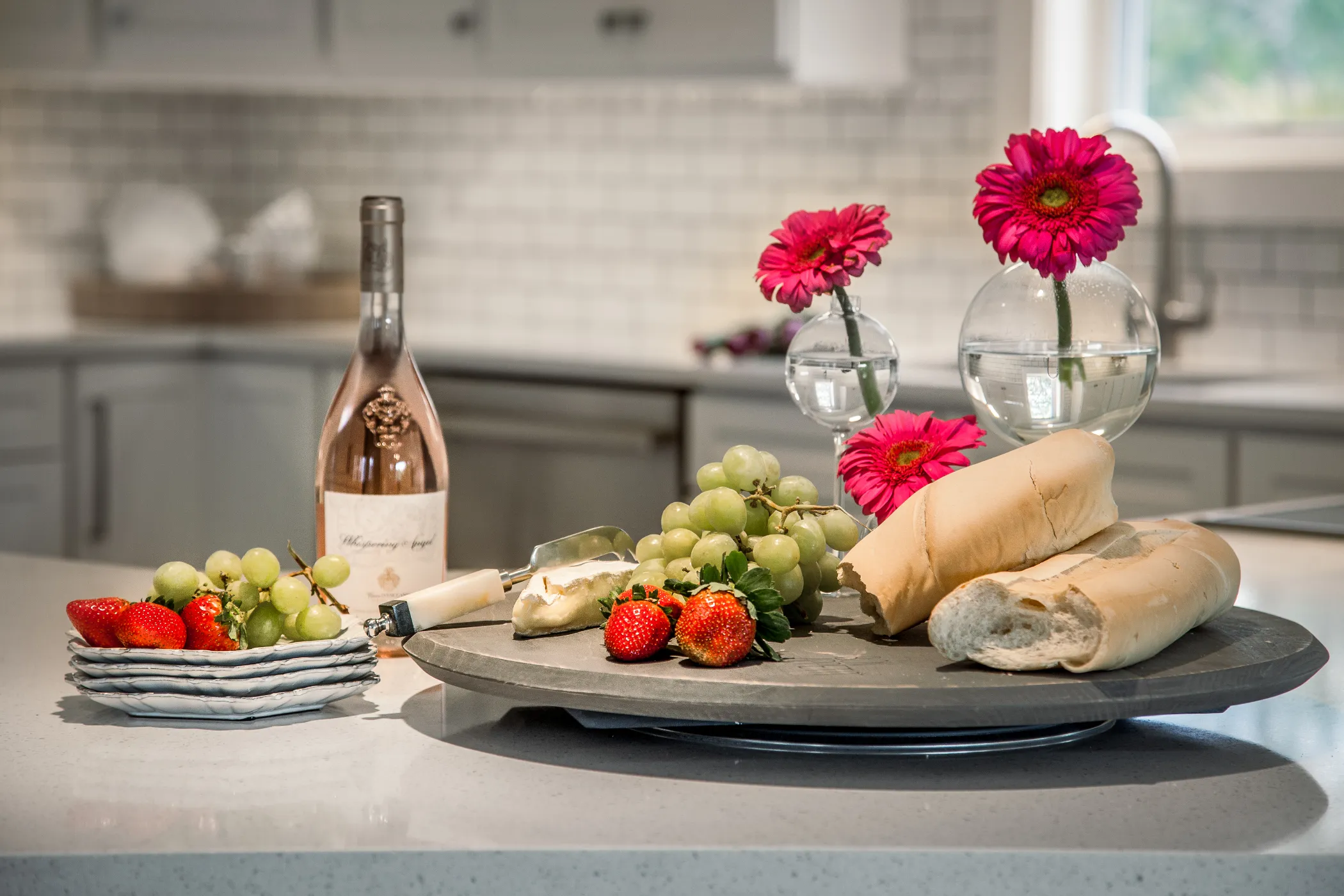 A kitchen counter with baguettes, strawberries, grapes, cheese, wine bottle, and pink flowers in vases.