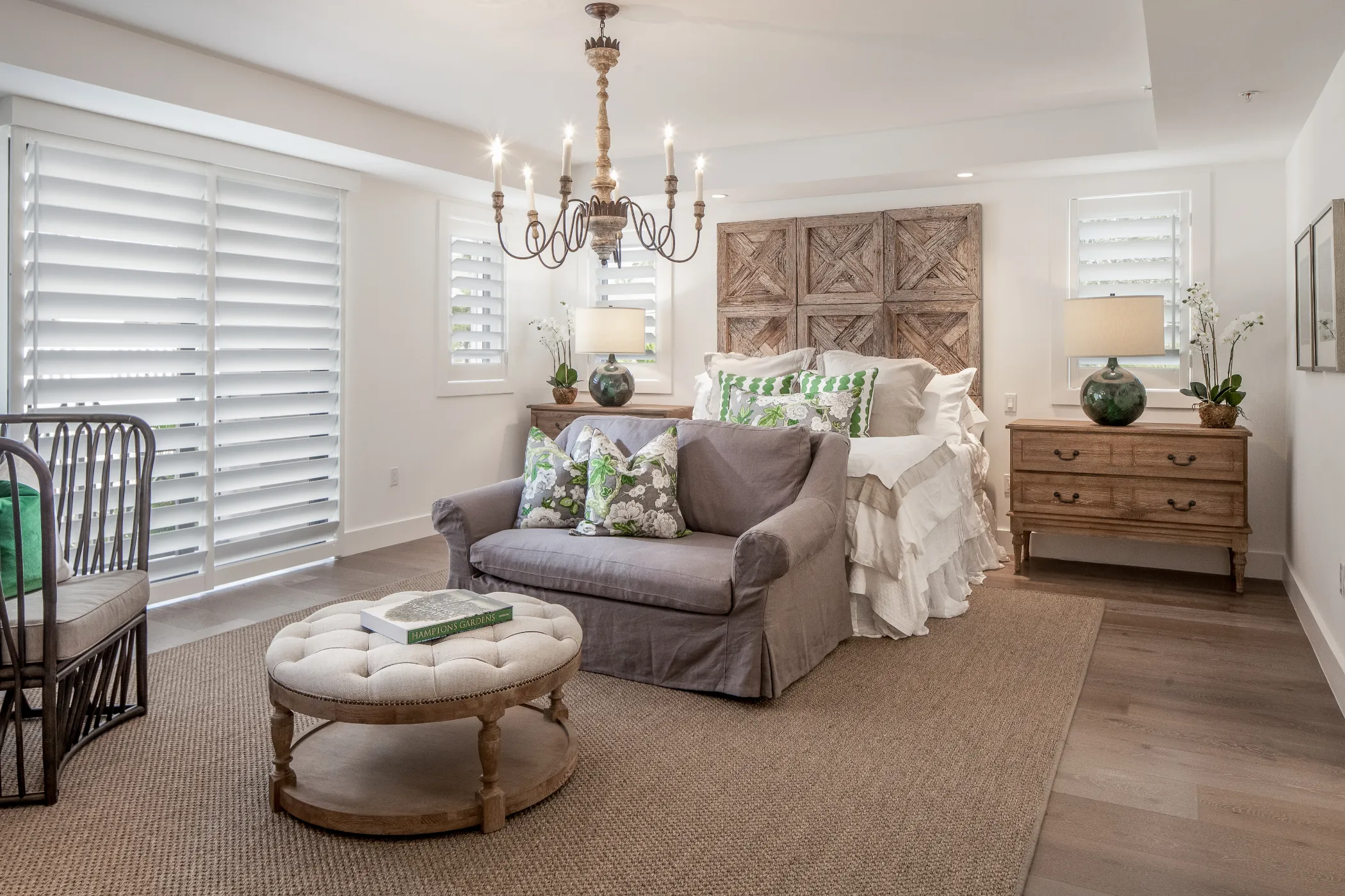 A cozy bedroom with a large bed, gray sofa, round ottoman, and wooden dresser under a chandelier.