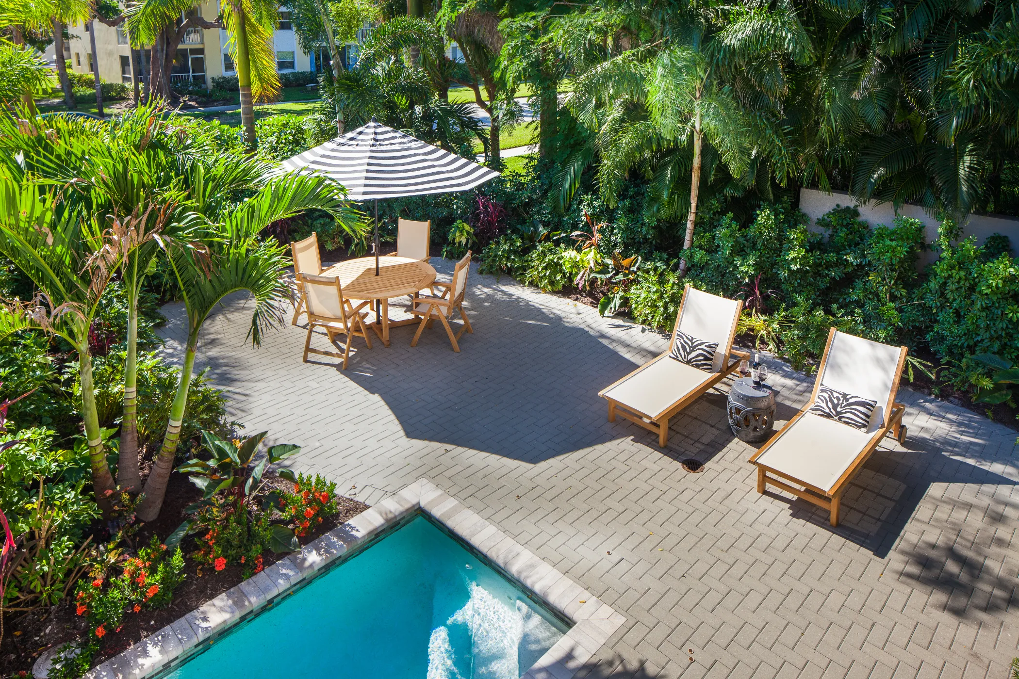 Outdoor patio with a round wooden table, umbrella, and two lounge chairs beside a small swimming pool.