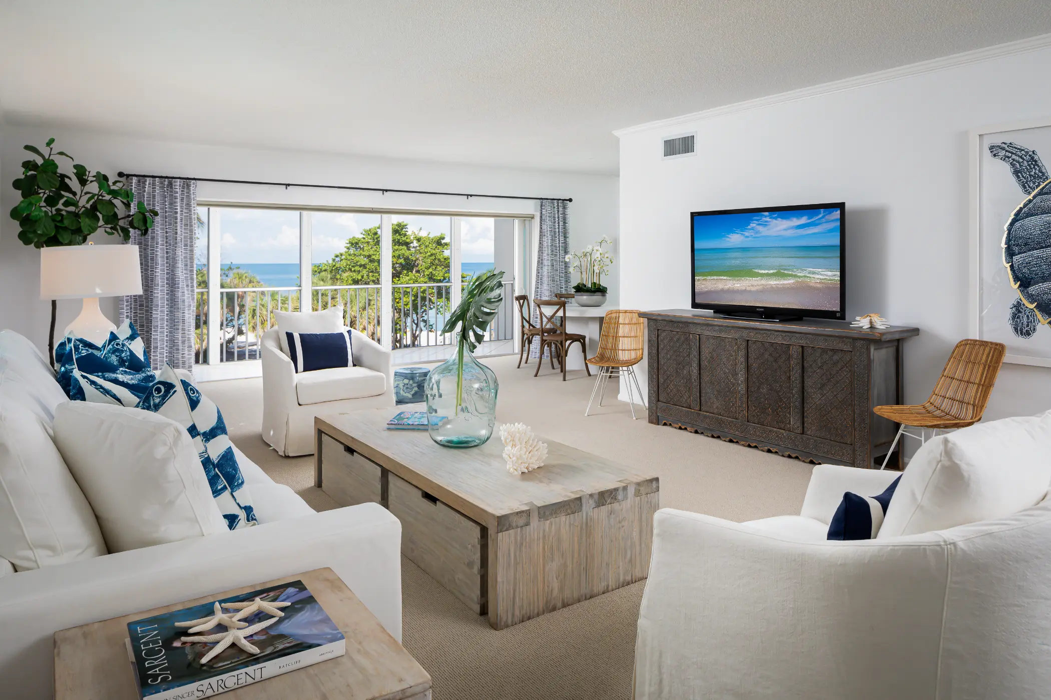 A modern living room with white sofas, a flat-screen TV, and large windows showing an ocean view.