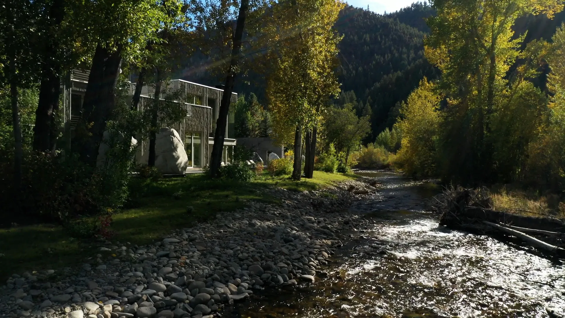 Modern cantilevered roof and stone facade house by architect Susan Desko, set along a forested riverbank in Sun Valley, Idaho, with sunlight streaming through the trees.