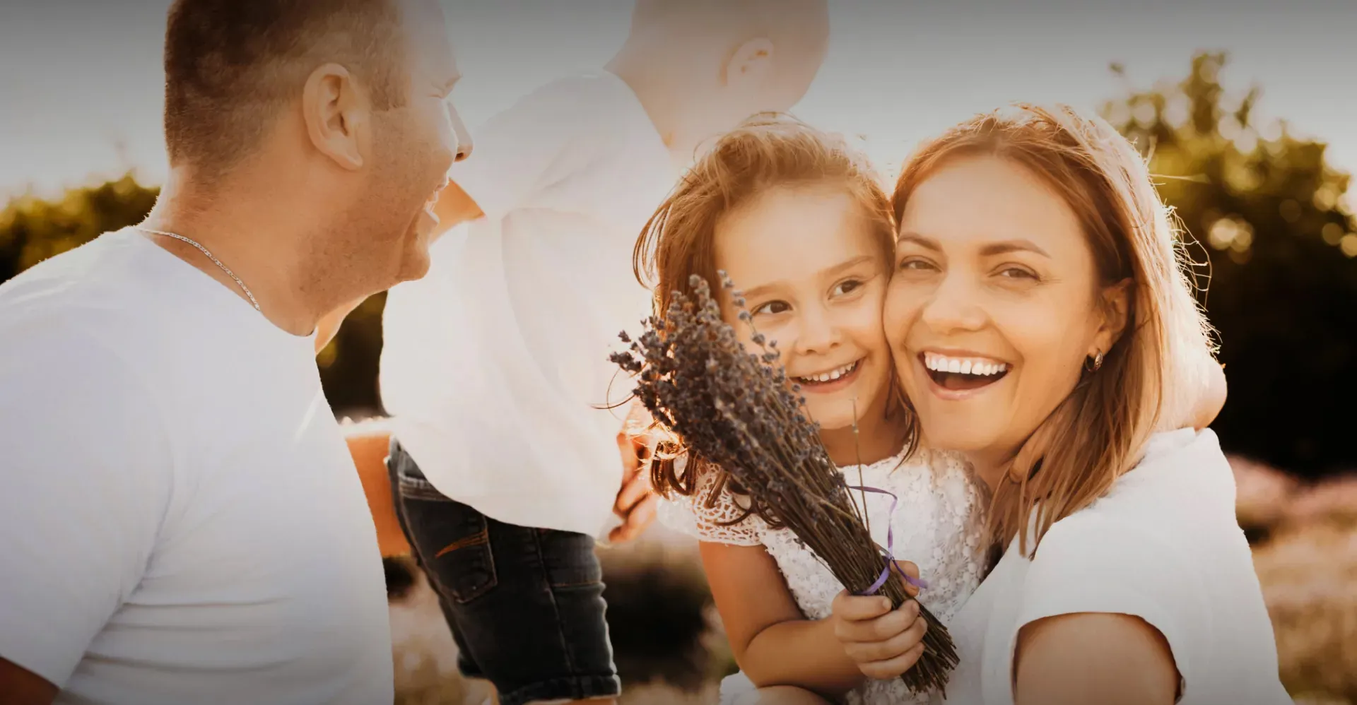 Smiling mother hugging daughter holding a bouquet of lavender with father and son in background outdoors at sunset.