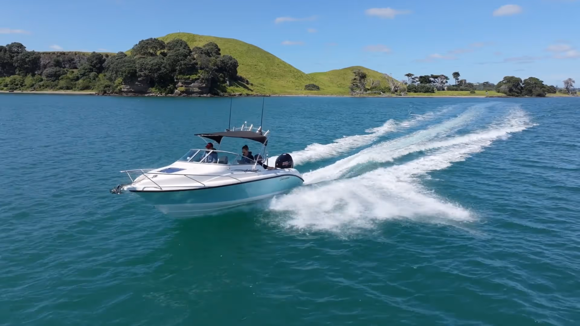 White motorboat speeding on blue water near green hills under a clear sky.