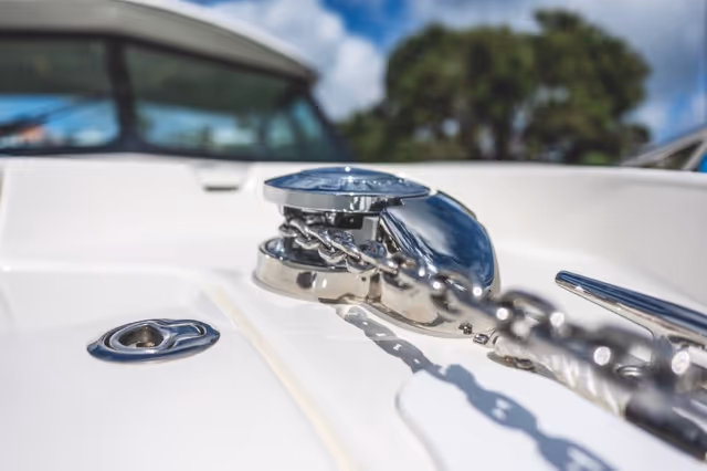 Close-up of a shiny metal anchor windlass and chain on the deck of a white boat with blurred trees and sky in the background.