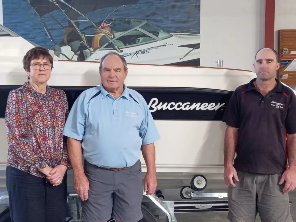 Three adults standing in front of a white boat with the name 'Buccaneer' displayed on its side.