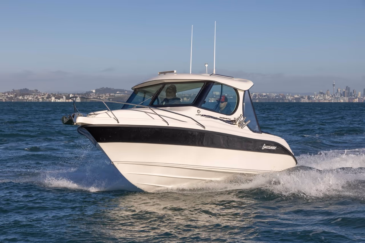 Small white and black motorboat named Buccaneer cruising on calm blue water with two people inside and a city skyline in the background.