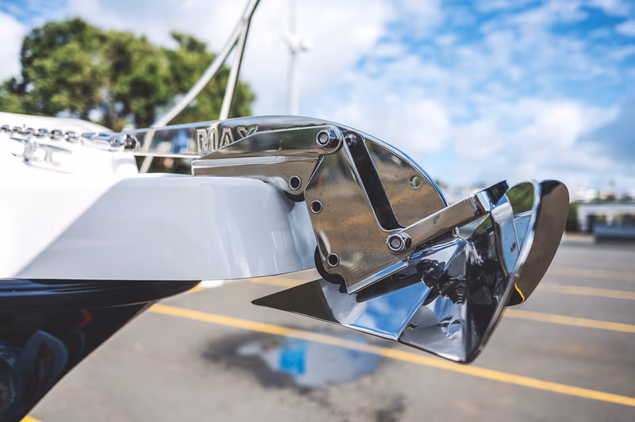 Close-up of a shiny stainless steel boat anchor mounted on a white boat hull with a blurred background of parking lot and trees.