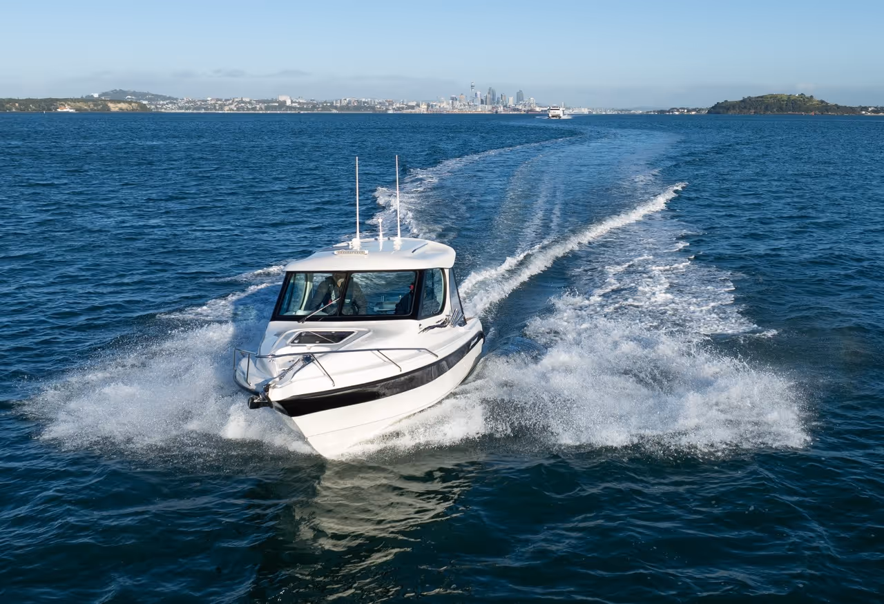 White motorboat speeding on blue water with a city skyline and islands in the distant background.