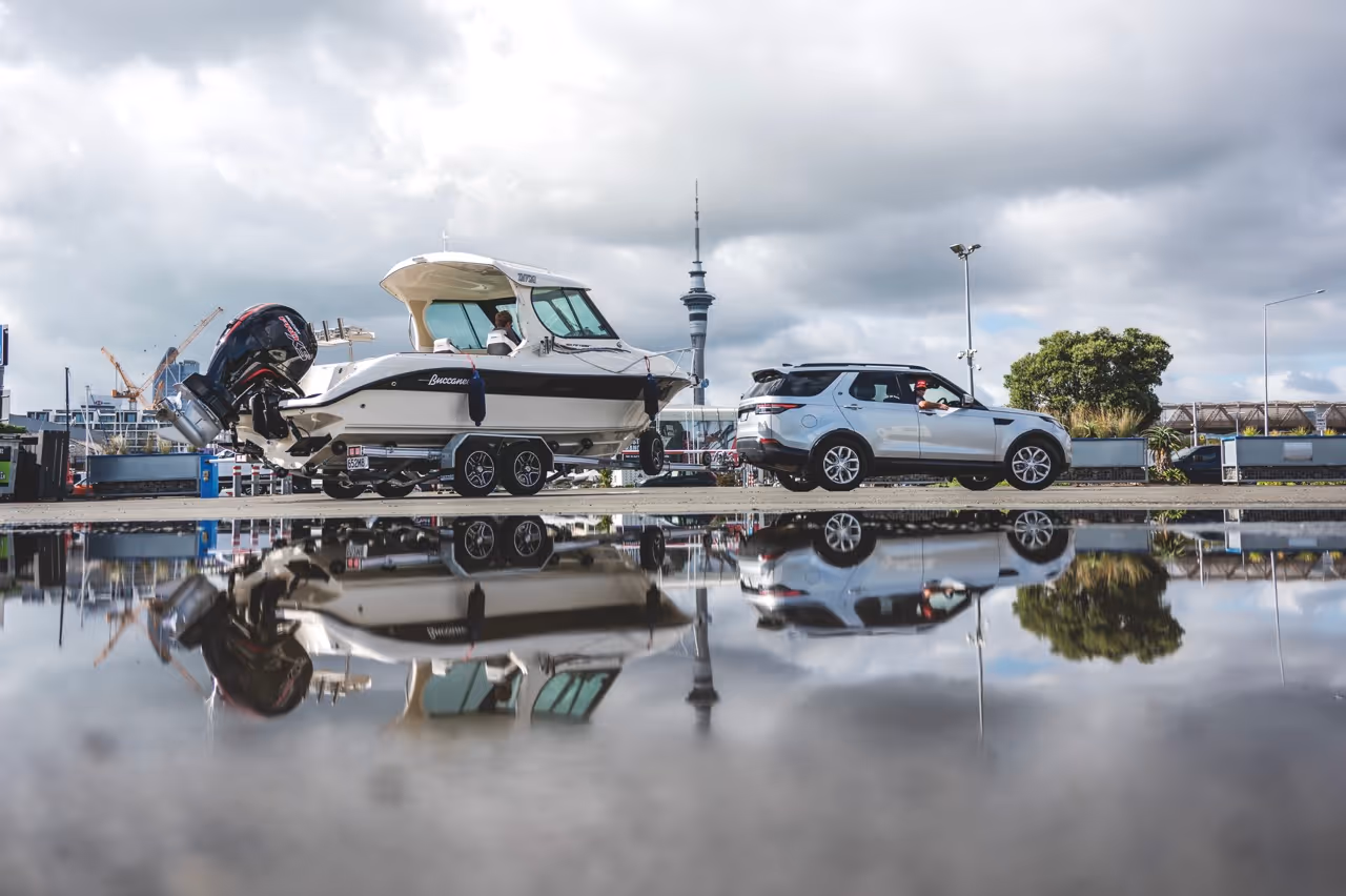 Silver SUV towing a white Buccaneer boat on a trailer, both reflected in a large puddle on a cloudy day near an urban area.