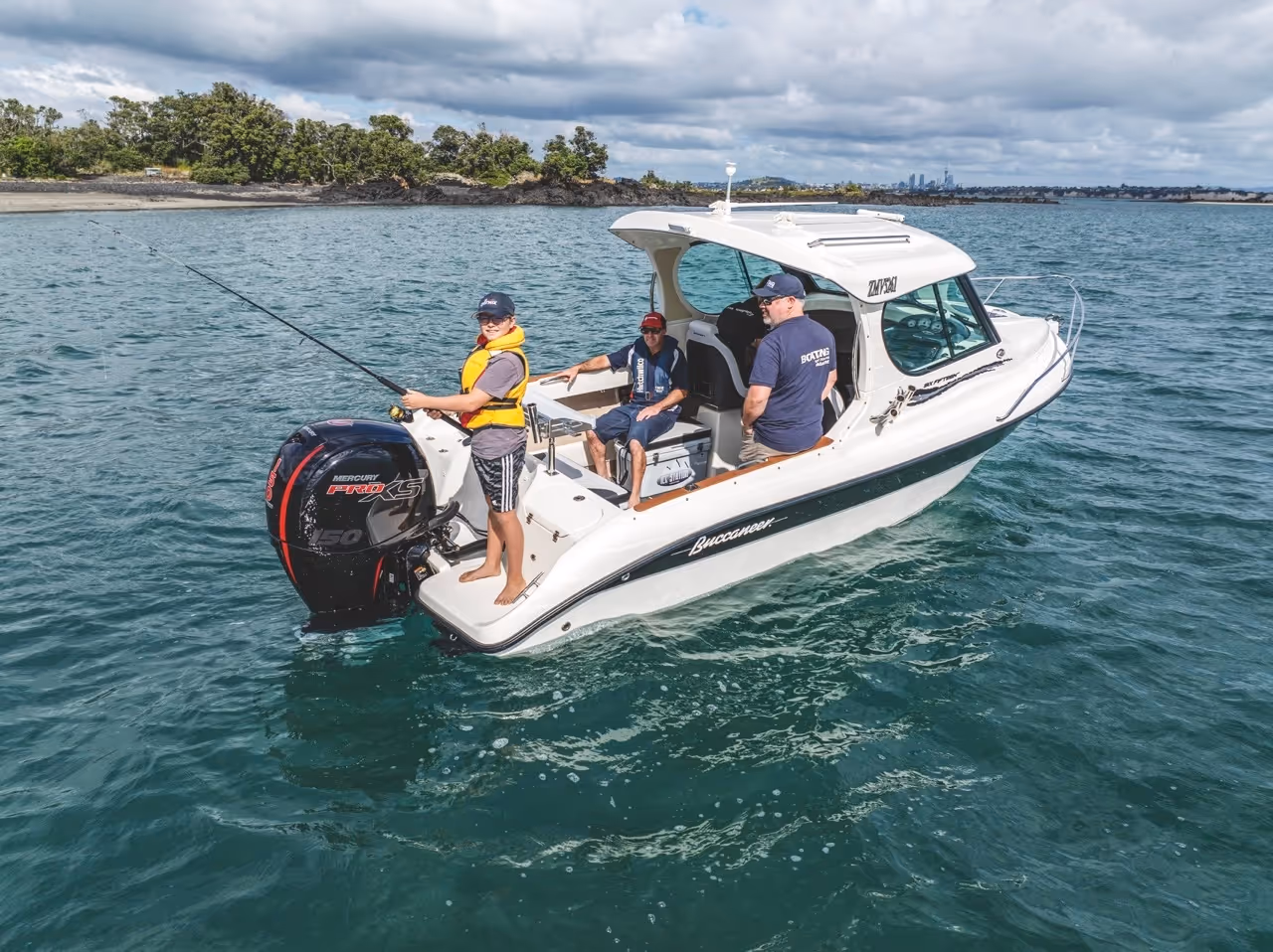 Three people on a white Buccaneer motorboat fishing in calm blue waters near a tree-lined shore under a cloudy sky.