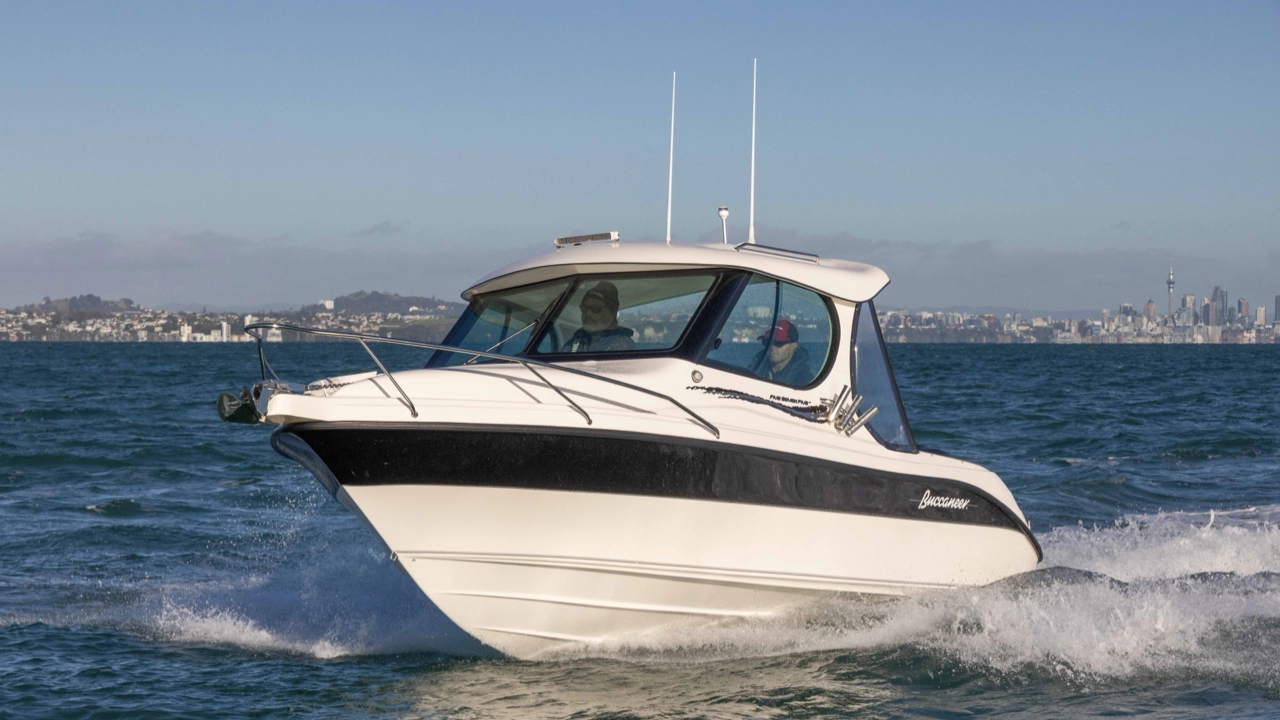 White and black Buccaneer motorboat cruising on blue water with two people inside and a city skyline in the background.