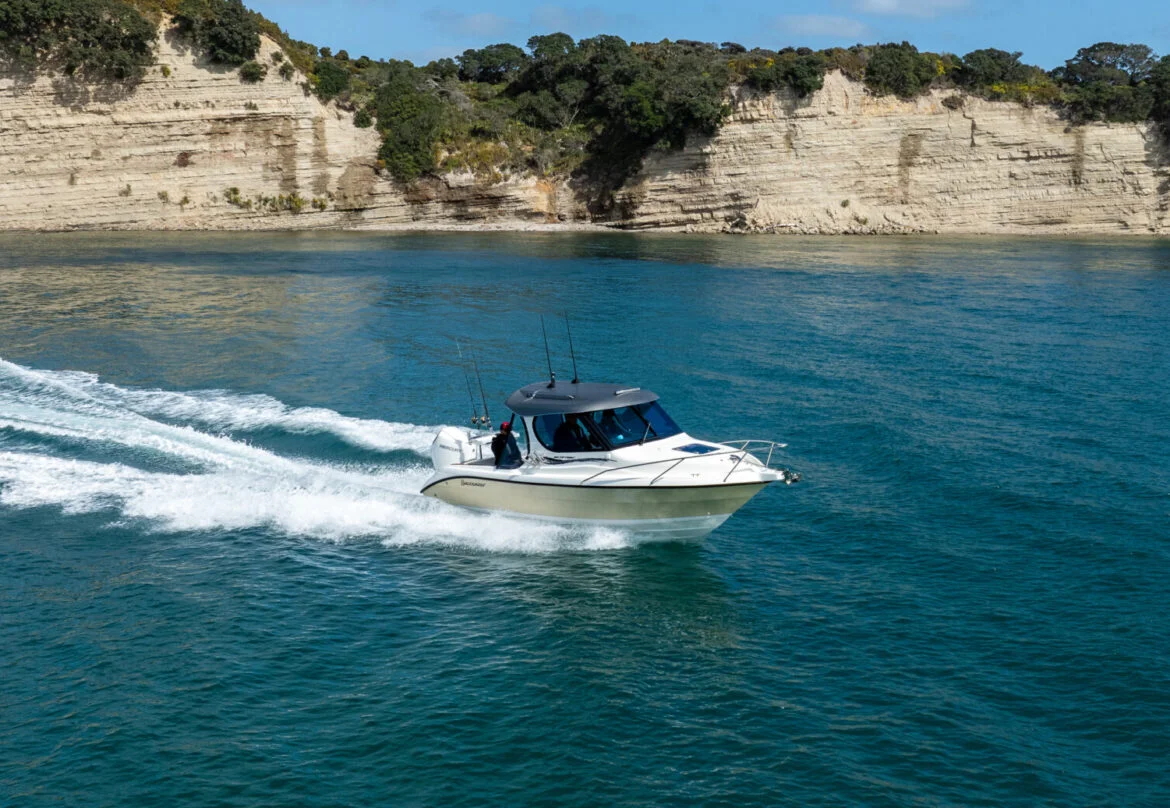 White motorboat with two people cruising on blue water near rocky cliffs with greenery under a clear sky.