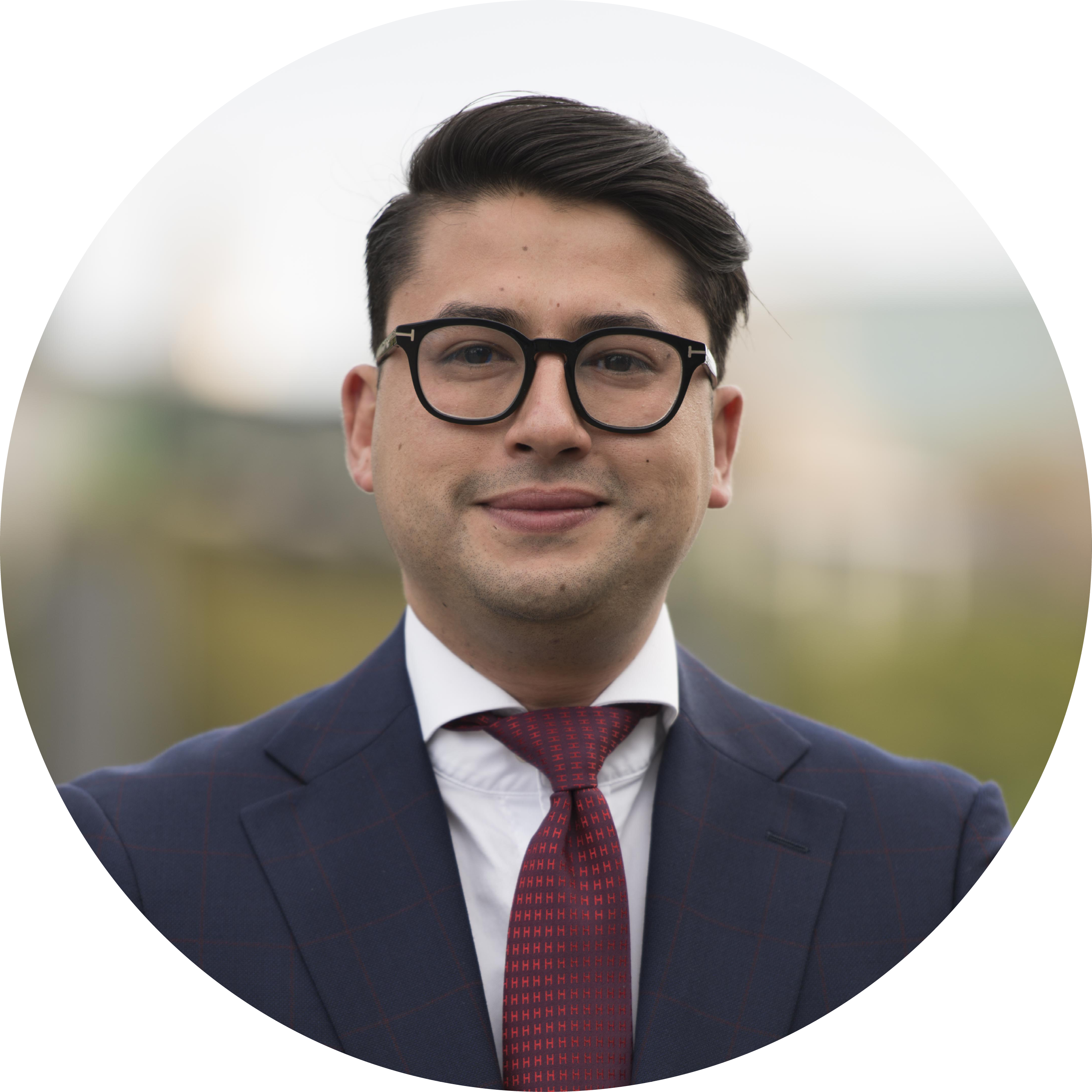 Man wearing black glasses, a dark blue suit jacket, white shirt, and a red tie, smiling slightly with blurred outdoor background.