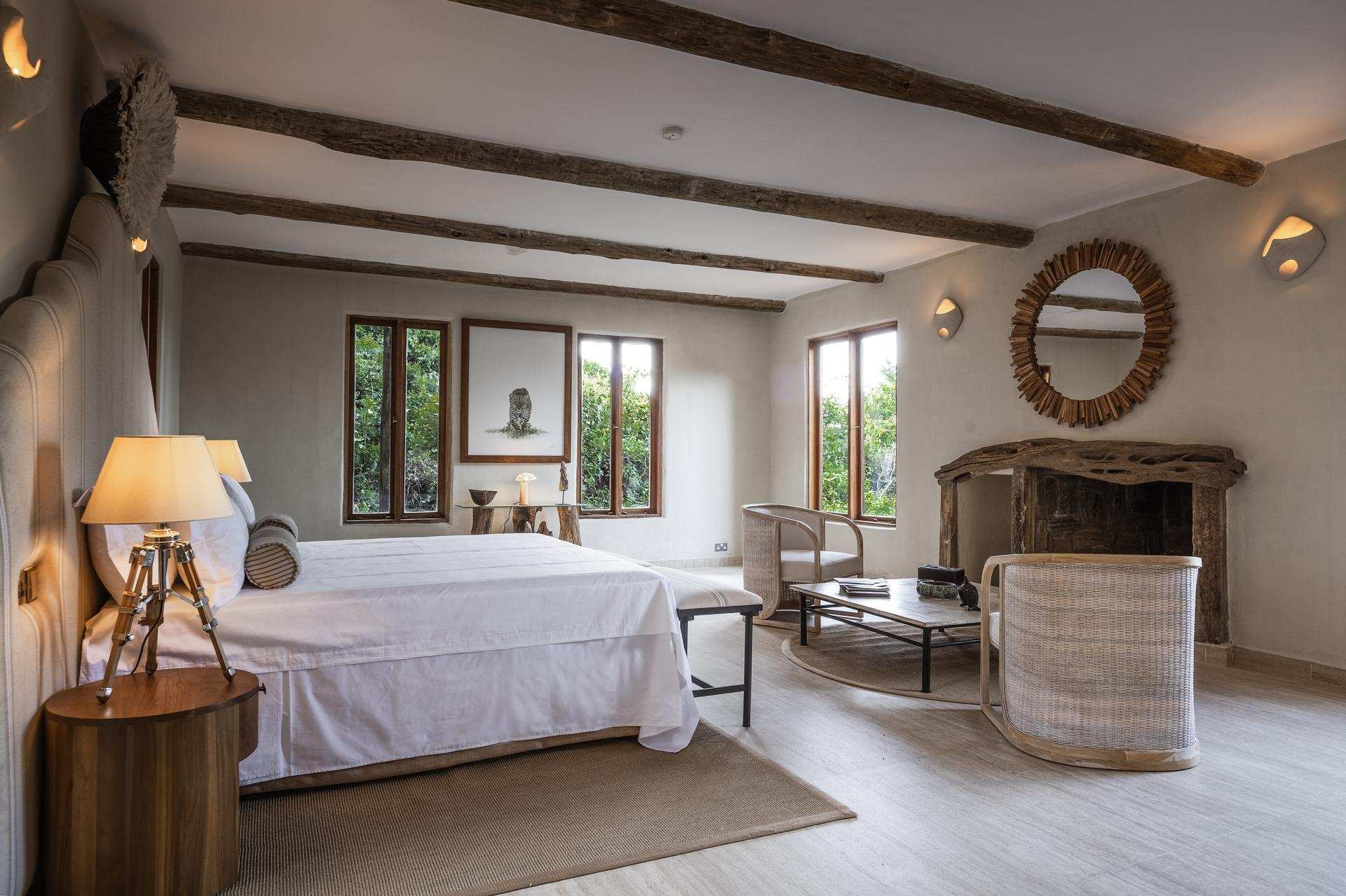 A spacious, sunlit bedroom with rustic wooden ceiling beams, a king-sized bed with white linens, natural wood furniture, and soft neutral tones. The room features a large mirror above a carved wooden fireplace and several windows overlooking greenery.