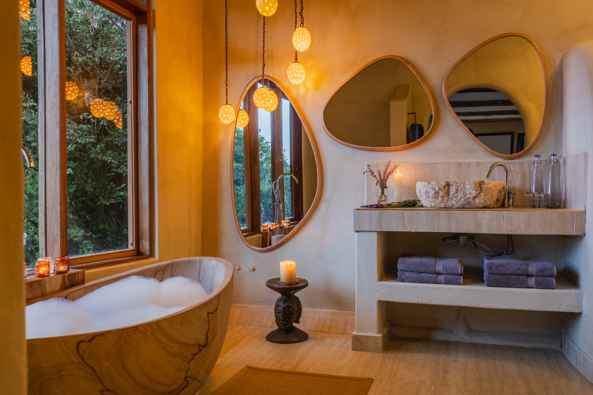 Luxurious bathroom with a carved stone bathtub by a large window overlooking greenery, softly lit by hanging pendant lights; stone sink with organic mirrors, candles, and natural decor creating a serene spa-like atmosphere.
