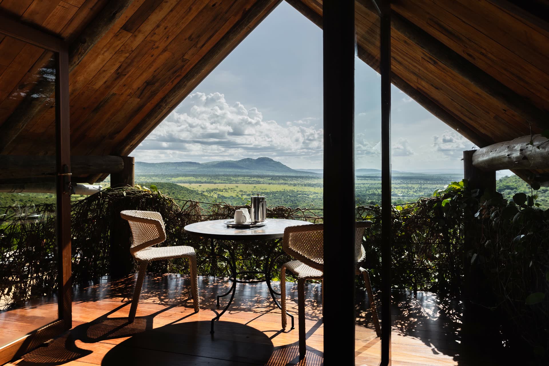 A cozy private balcony under a pitched wooden roof, featuring a small round table and two woven chairs overlooking a vast savannah landscape with rolling hills under a bright, cloud-dappled sky.