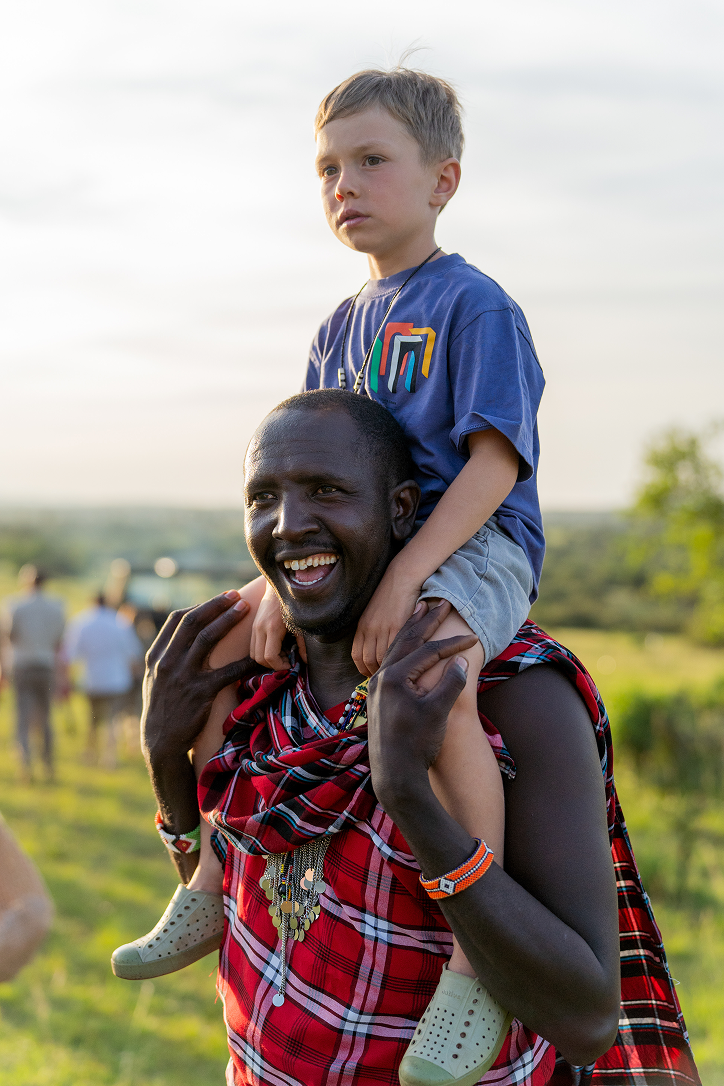 Young safari guest riding on a Maasai guide’s shoulders during a family safari experience.