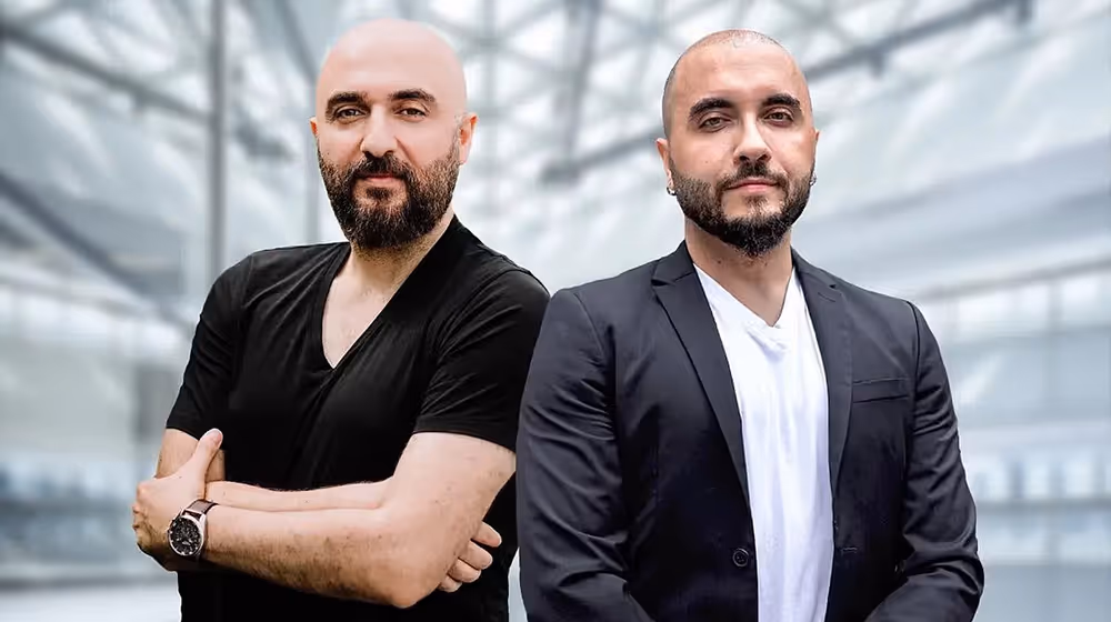 Two men with shaved heads and black suits stand side by side.