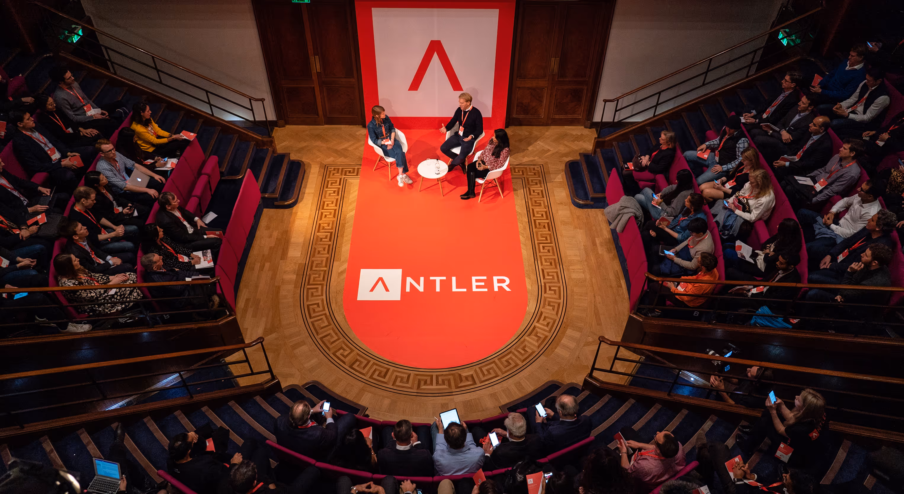 A group of people sitting in a large room with a red carpet.