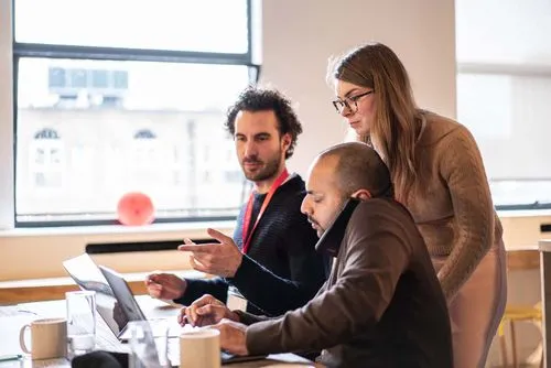 Three coworkers collaborating at a table, with one person on a phone call while the others review something on a laptop.