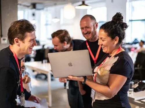 A small group of colleagues gathered around a laptop, laughing and discussing something on the screen in an open office space.
