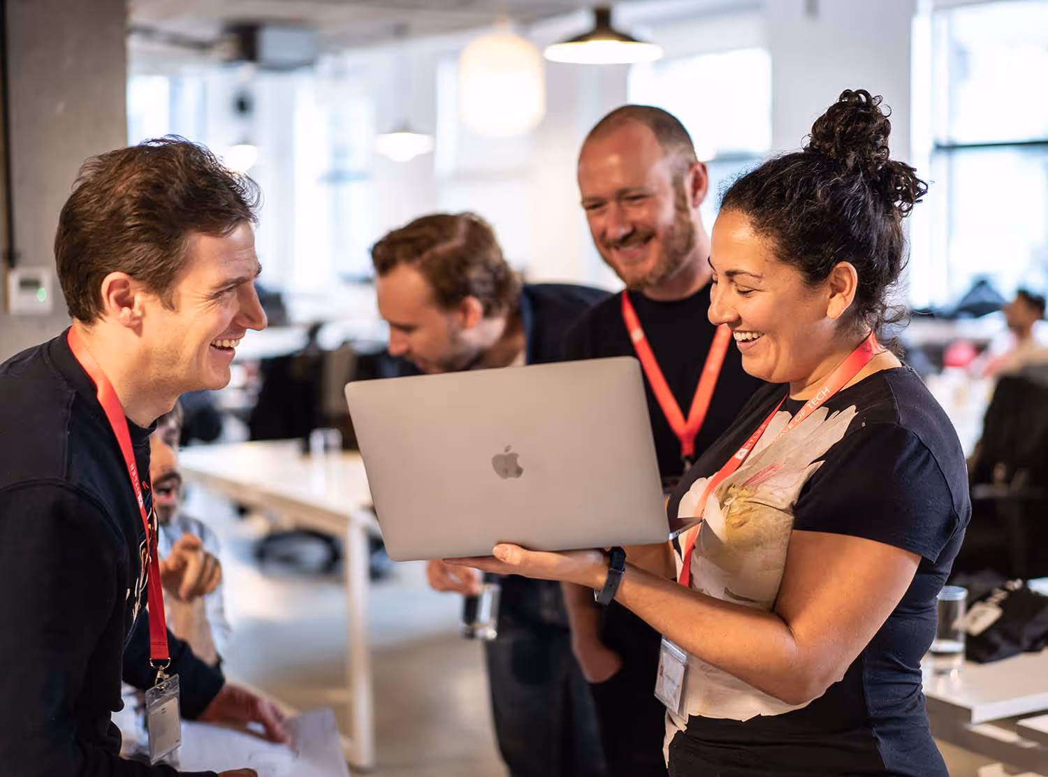 Um pequeno grupo de colegas reuniu-se em torno de um laptop, rindo e discutindo algo na tela em um escritório aberto.