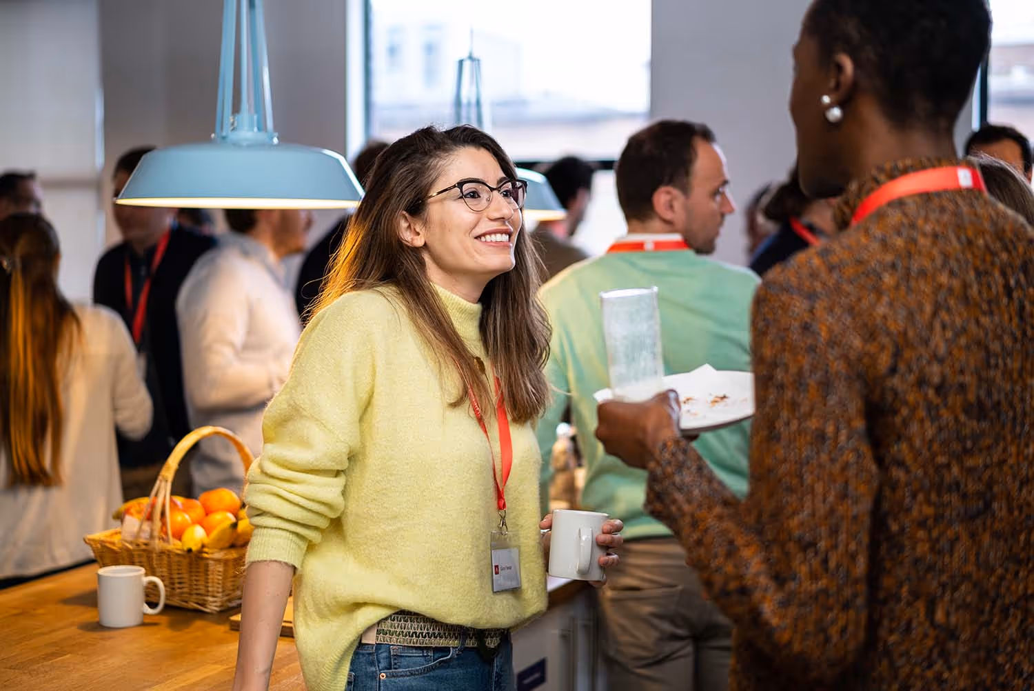 A woman smiling and talking with another attendee during a casual networking event in a bright, modern office kitchen.
