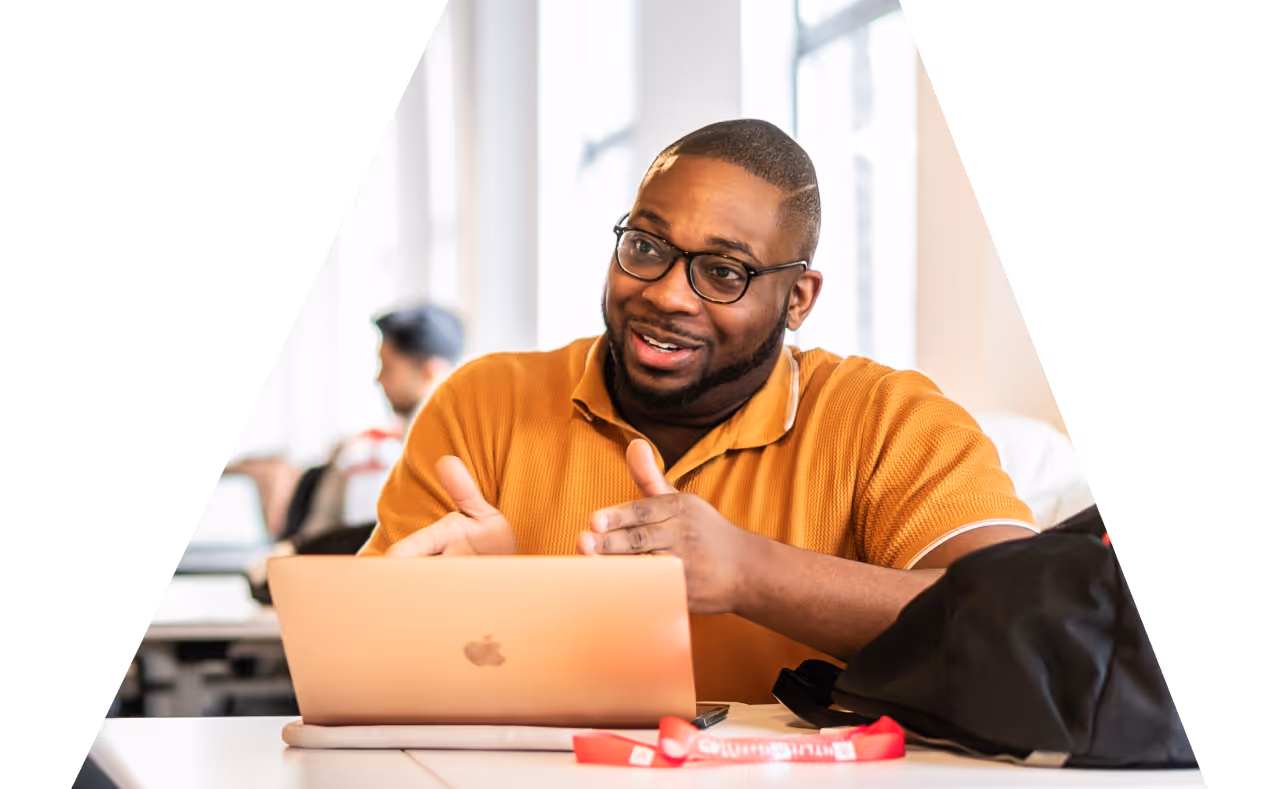 A man in a mustard-colored shirt speaks while gesturing, seated at a laptop during a group discussion.