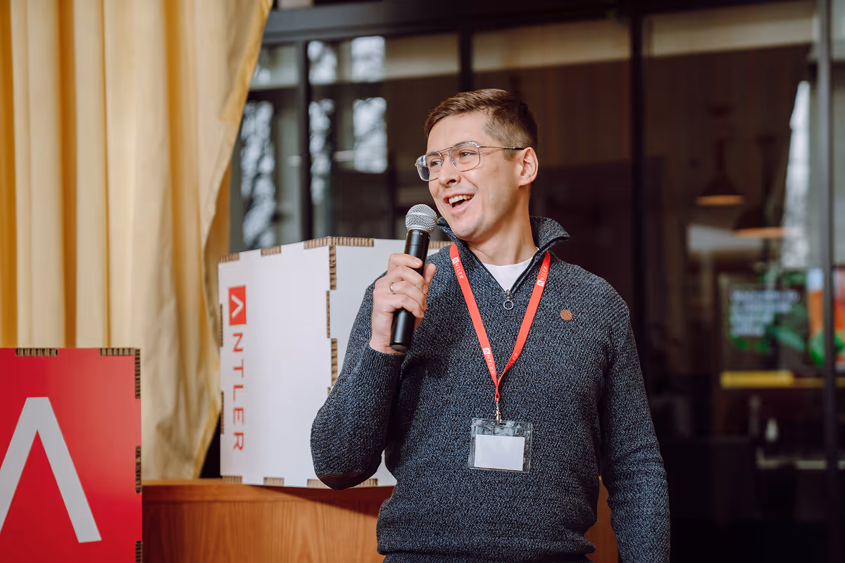 A person holding a microphone and smiling while speaking at an indoor event. They are wearing glasses, a dark sweater, and a red lanyard. Branded Antler signage is visible in the background.