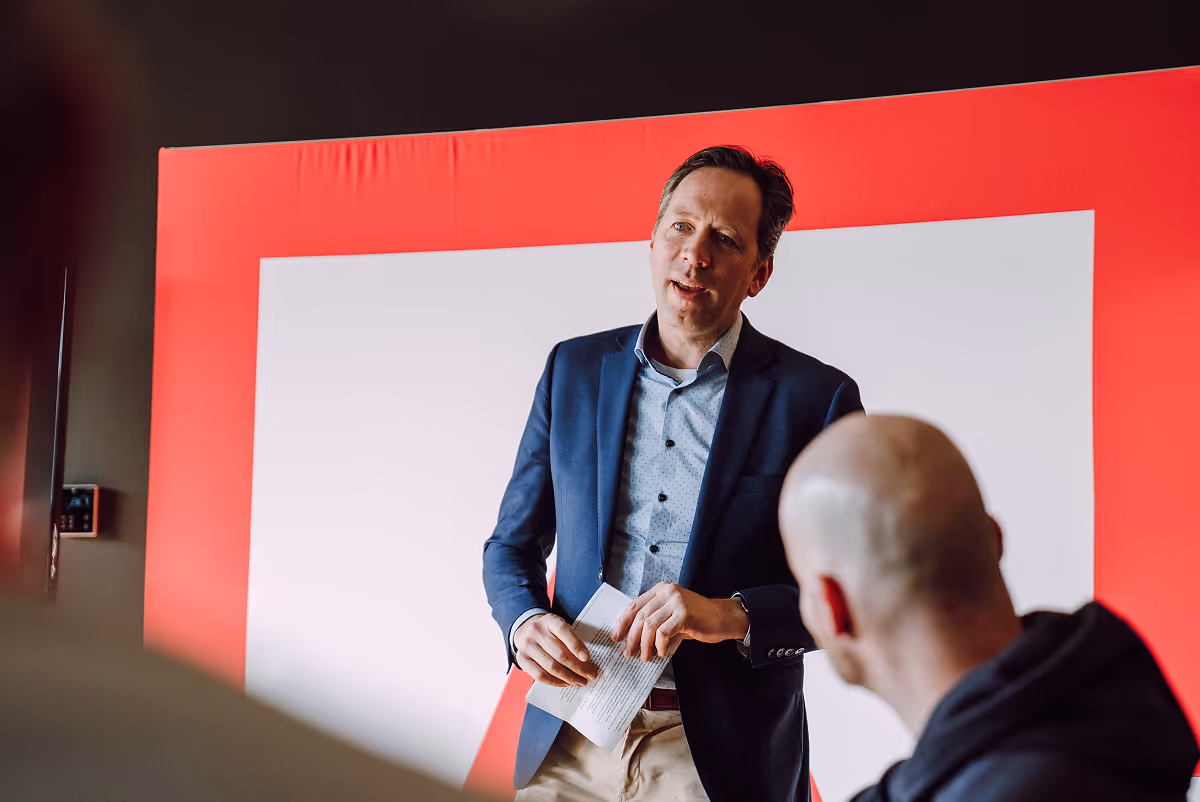 A man in a blue blazer speaks while holding notes, standing in front of a red-and-white backdrop, addressing a small seated group in a casual meeting setting.