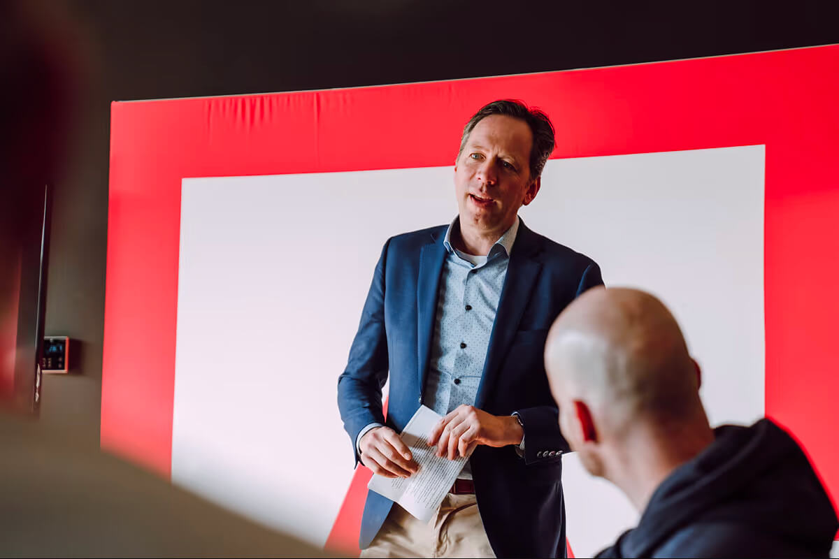 Man in a blue blazer holding papers and speaking to an audience against a red and white background.