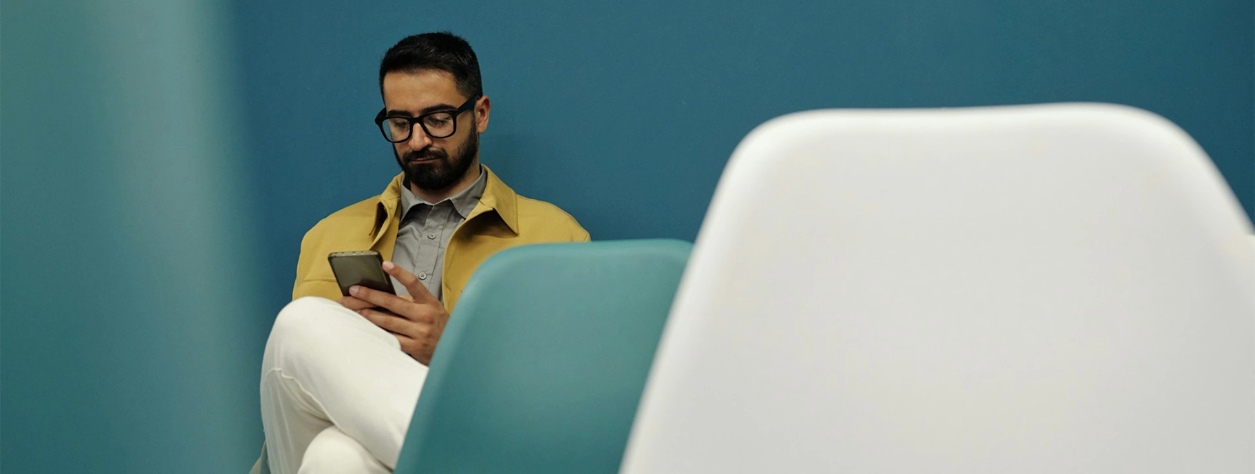 Man with glasses sitting on a chair against a blue wall, looking at his smartphone.
