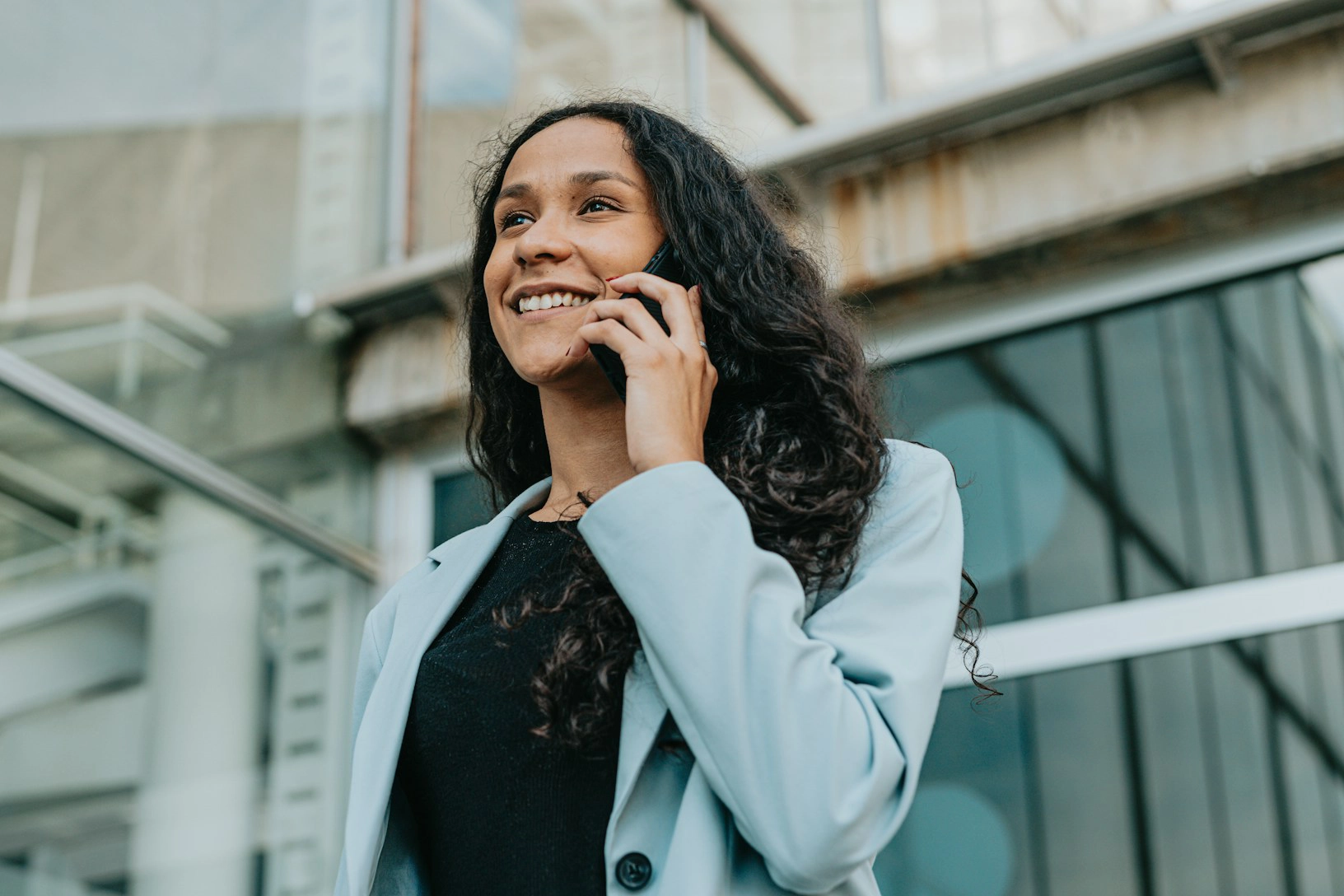 Smiling woman with long curly hair wearing a light blue blazer, talking on a smartphone outside a modern building.