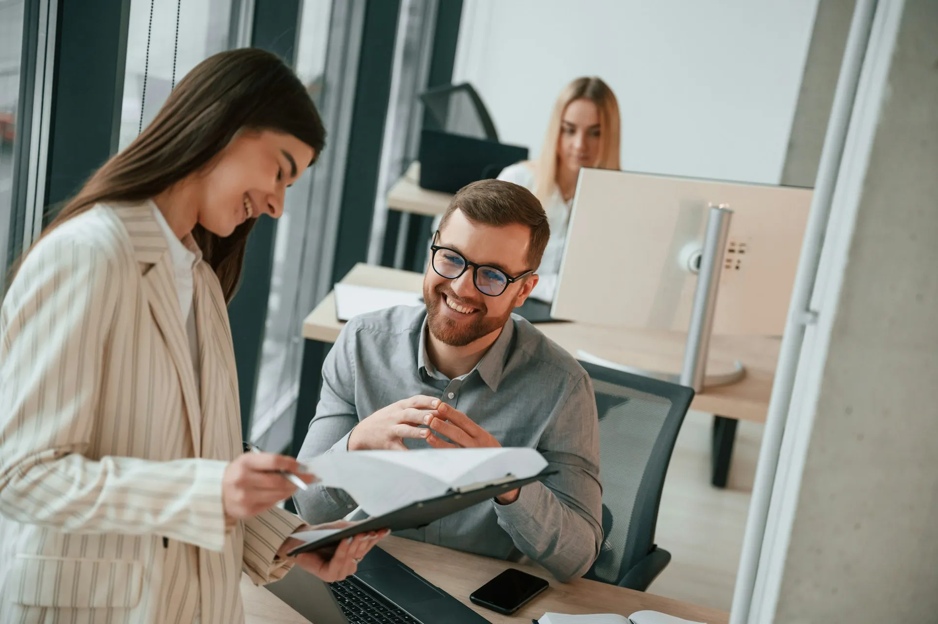 A woman shows a document to a smiling man, both collaborating in a modern office. Another woman works in the background.