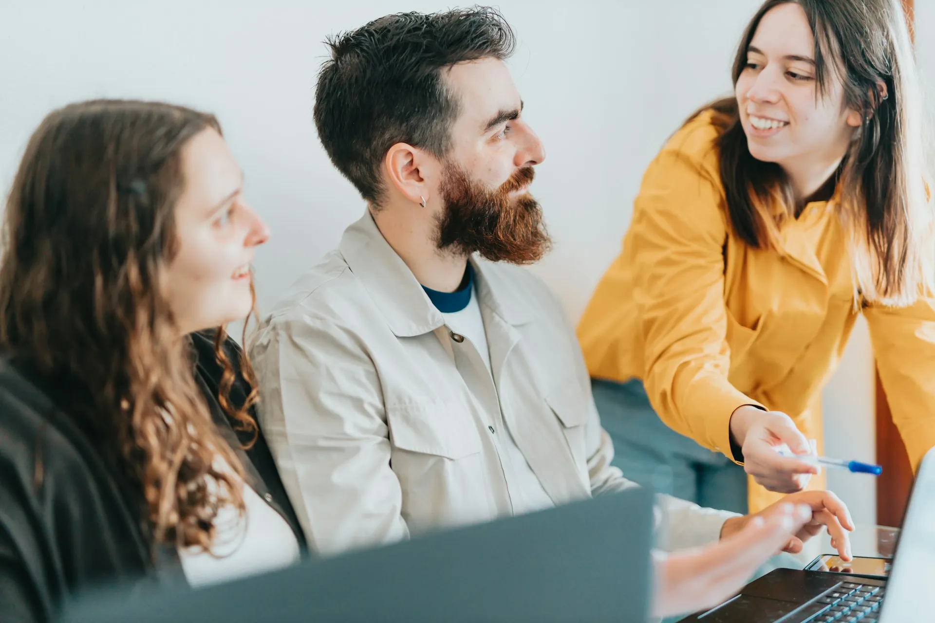 Three young adults engaged in a discussion around a laptop, with one woman pointing at the screen with a pen.